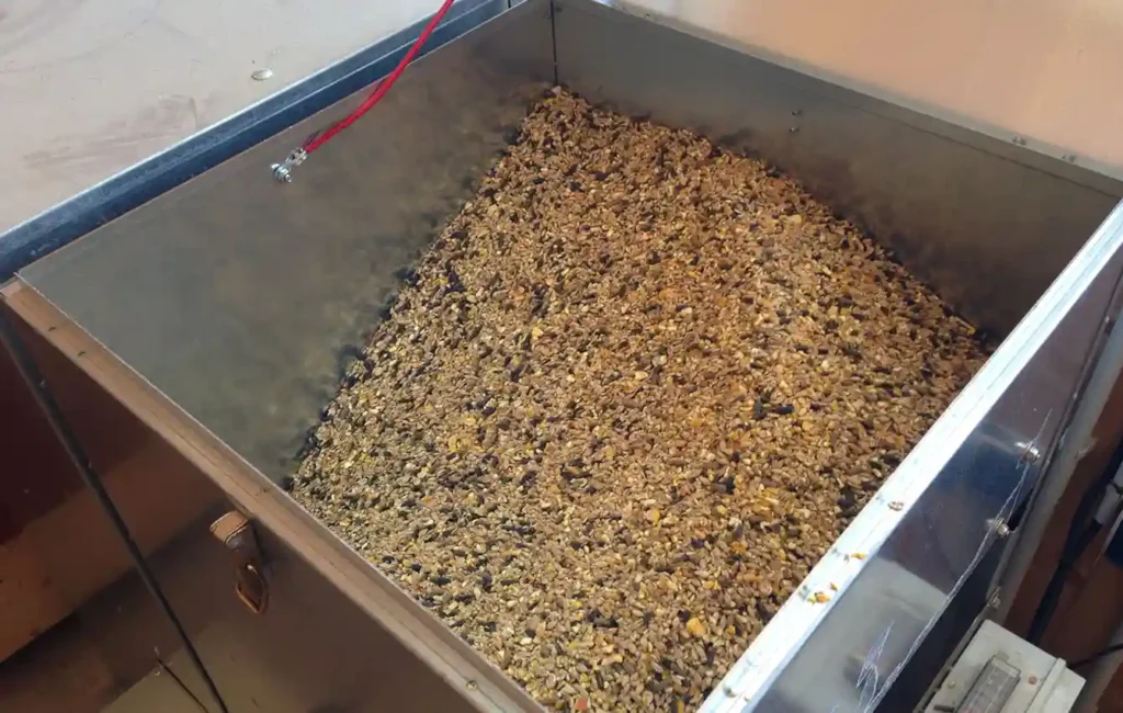 Close-up of a light-colored feed mixture (concentrated feed) for horses: You can see a coarse structure with light brown pellets mixed with golden yellow cereal flakes and small, darker grains. The individual components are clearly visible, and the light highlights the texture.  Dosing container for HIT concentrated feed stations with a filling volume of 150 liters