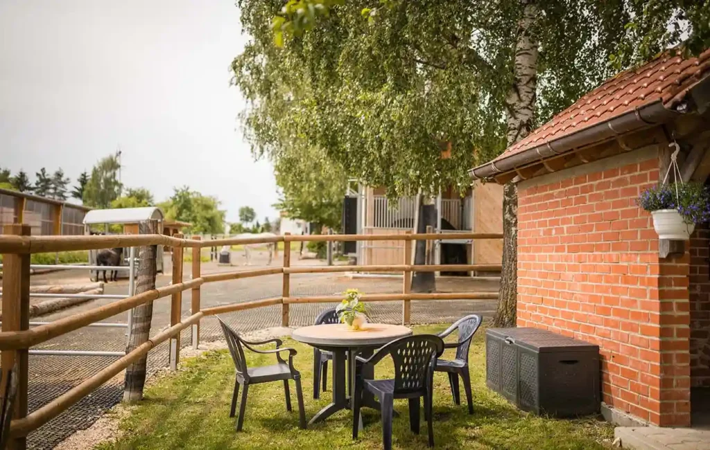 A small round garden table with four chairs stands on a green lawn. To the right, part of a building can be seen, surrounded by a fence. In the background are stables with horse boxes and horses grazing. The scene appears calm and inviting, like a cozy place to linger near the horse stable.    In the HIT-Active Stable concept, a capital “F” stands for “Freetime”