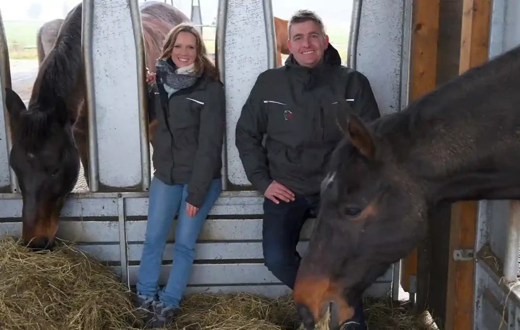 The picture shows the inside of a horse stall. Horses standing outside stick their heads through two separate openings to eat hay. Inside the stall, a woman sits on the edge of the left opening and a man sits on the edge of the right opening. Both smile at the camera while the horses calmly eat from the openings next to them. A large “F” in the HIT active stable concept stands for “focus.”