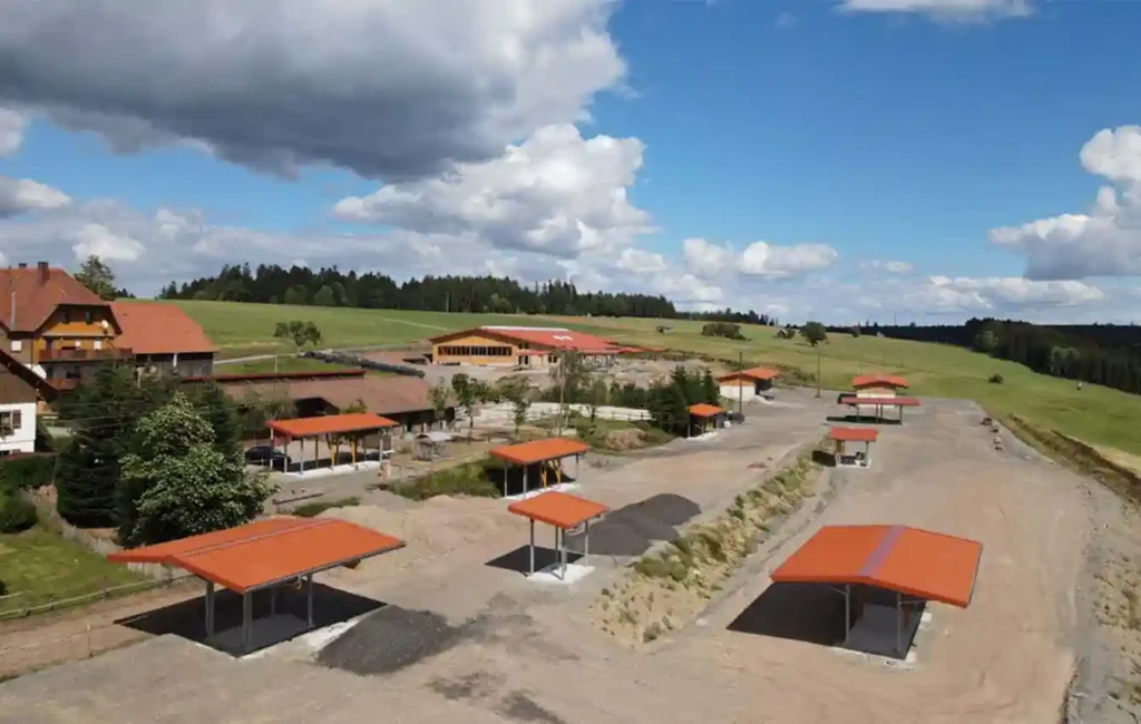 The image shows a farmyard from a bird's eye view. Several small shelters or open shelters are scattered across the grounds. There is a larger house on the left-hand side. The entire area is surrounded by green meadows and trees and is located on a slight slope. The photo provides a comprehensive overview of the farm and its outdoor areas. The principle of the HIT active stable: Spatial separation of the functional areas marketplace (lying area, automatic feeders, racks, drinking troughs) and structured walkways or trails including rolling area, deadwood hedge, horse stairs, tree trunk maze, water ford, etc.