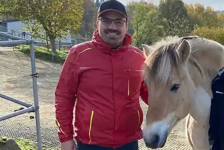 Contact person 7 In the center of the picture stands a smiling man looking directly into the camera. Next to him stands a light-colored horse. Both are located on sandy ground that slopes slightly. There are trees in the background.