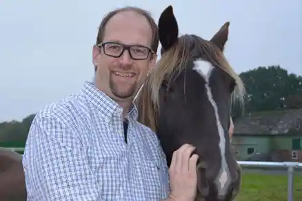 Contact person 8 A man holds the head of a black horse positioned to his right and smiles. Only the head of each of them is visible. A barn and a meadow are faintly visible in the background, but the bright sky dominates the scene.