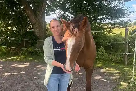 Contact person 3 A woman stands to the left of a brown horse and holds her hands to the animal's mouth. Behind them runs a fence separating the trees behind them from the sandy ground on which the woman and the horse are standing.