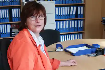 Contact person 2 A woman is sitting at her workplace on the left side of the picture and looking into the camera. Behind her are several shelves filled with folders.