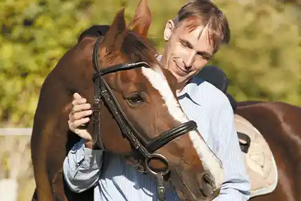 Contact person 9 A man holds the head of a brown horse standing to his left and smiles at the animal. The background is blurred, but shows green trees and natural vegetation.